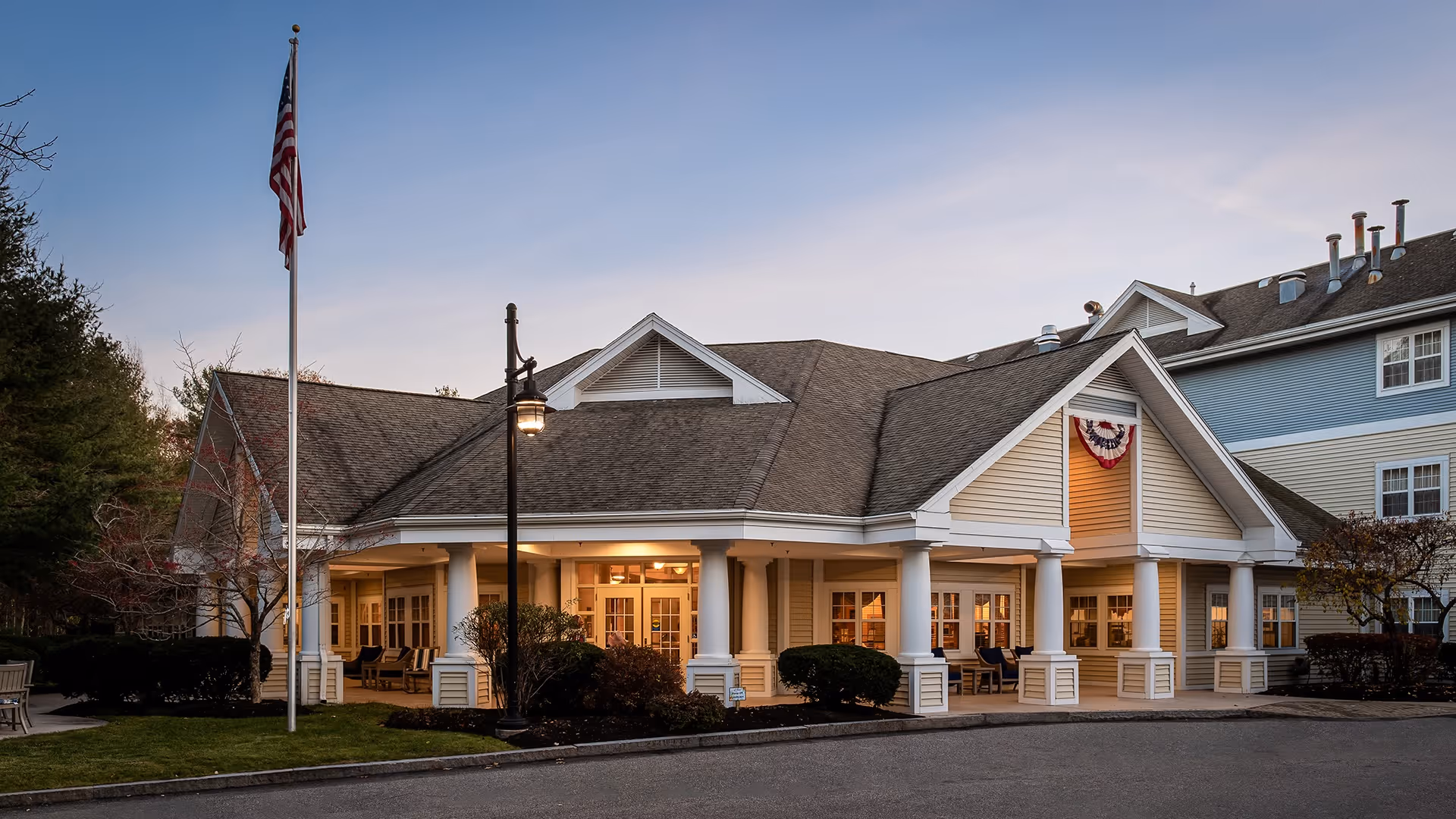 Exterior view of a senior living facility building with a covered porch supported by white columns, an American flag on a flagpole, and a street lamp. The building has beige siding with white trim and a gabled roof. There are bushes and trees around the building and the sky is clear with soft evening light.