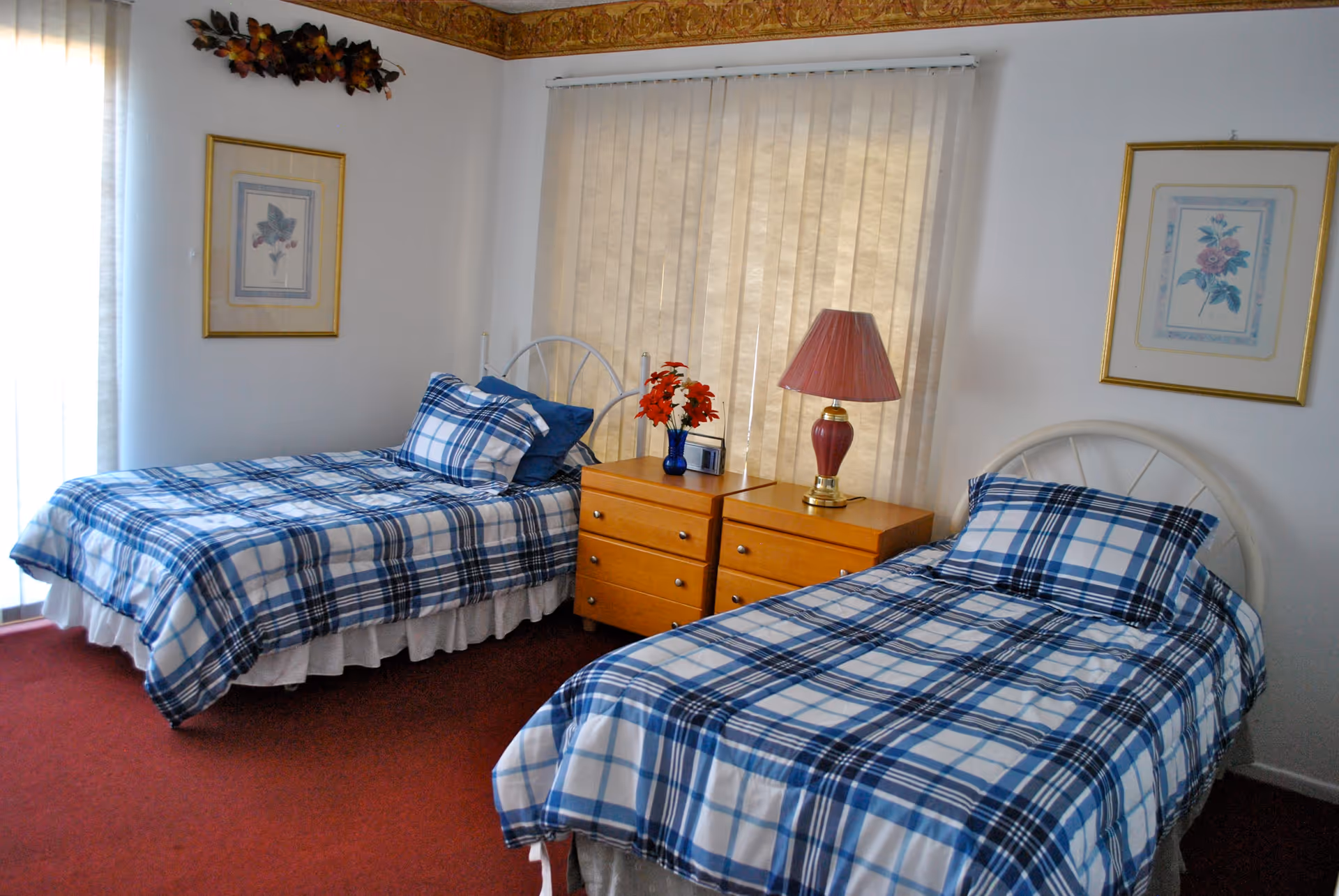 A bedroom with two single beds featuring blue and white plaid bedding. Between the beds is a wooden nightstand with a red lamp and a small vase with red flowers. The room has white walls with framed floral artwork and a window covered by beige vertical blinds. The floor is carpeted in red.
