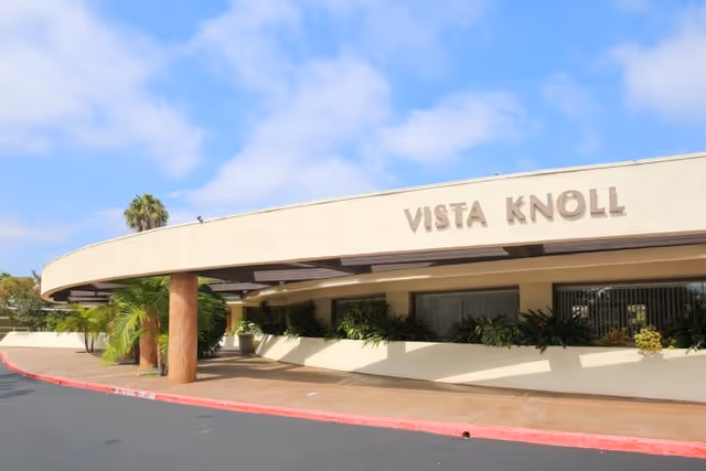Front entrance of Vista Knoll with a curved canopy, landscaping, and the facility name on the facade under a blue sky.