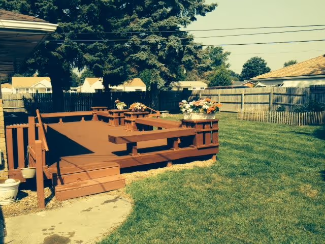 A backyard area with a wooden deck painted brown, featuring built-in benches and flower pots with colorful flowers. The deck is surrounded by a green lawn and enclosed by a wooden fence. Trees and neighboring houses are visible in the background under a clear sky.