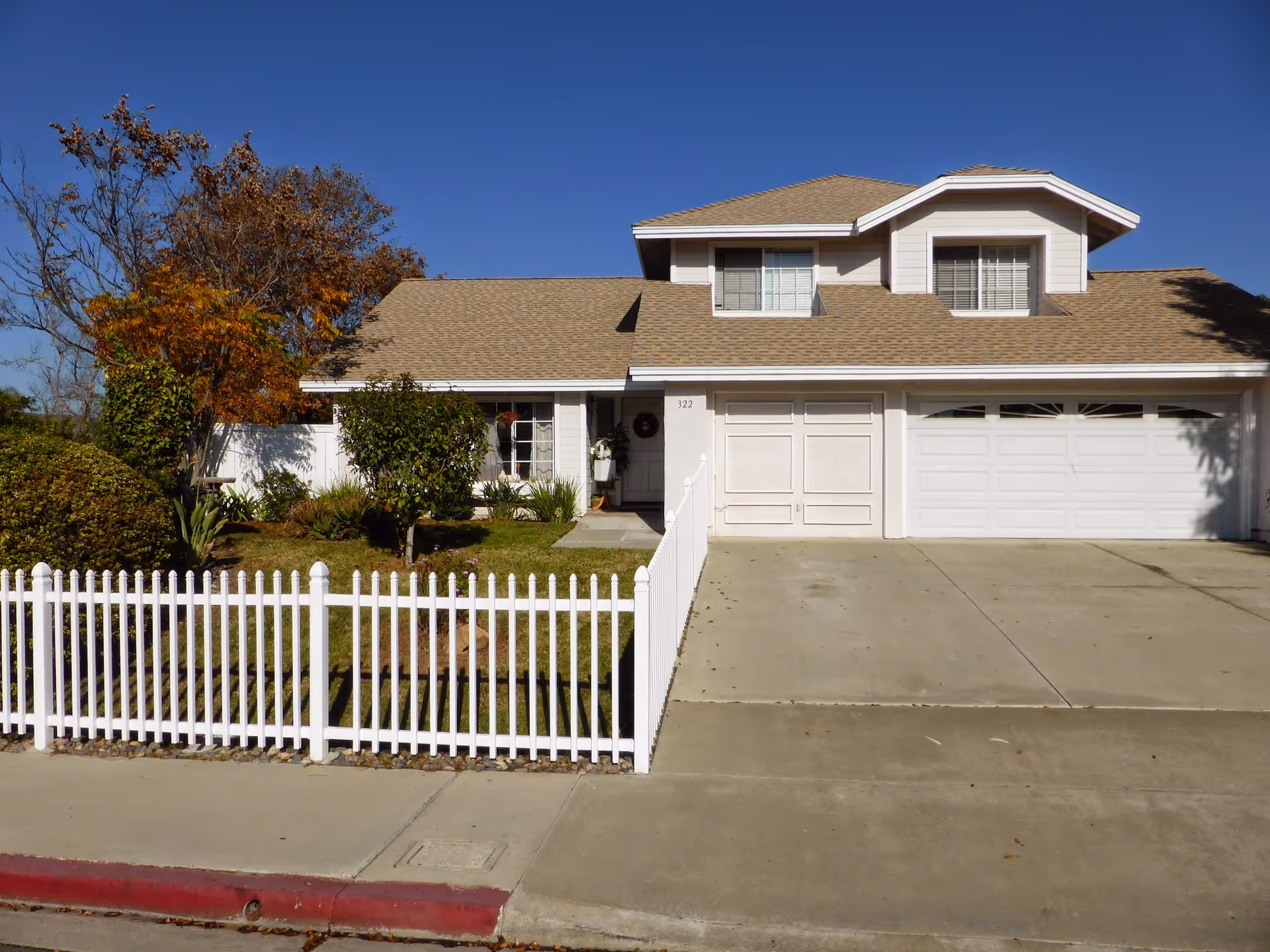 A two-story house with a beige roof and white exterior walls. The house has a white picket fence in front, a driveway leading to a double garage, and some trees and bushes in the front yard. The sky is clear and blue.