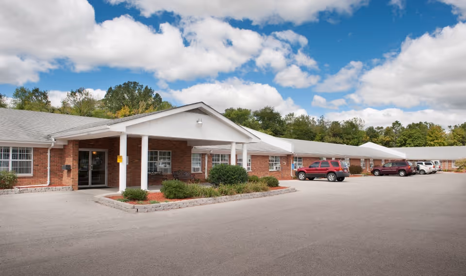 Exterior view of a single-story brick building with a covered entrance supported by white columns. Several cars are parked in front of the building, and there are trees and greenery in the background under a partly cloudy blue sky.