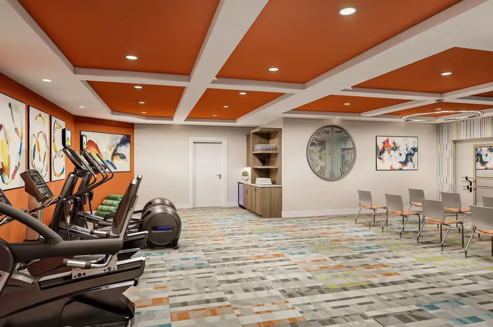 A fitness room with several elliptical machines lined up on the left side, colorful abstract paintings on the walls, a large round clock on the far wall, and a row of chairs arranged on the right side. The ceiling has orange recessed panels with white beams, and the floor is covered with a patterned carpet in various colors.