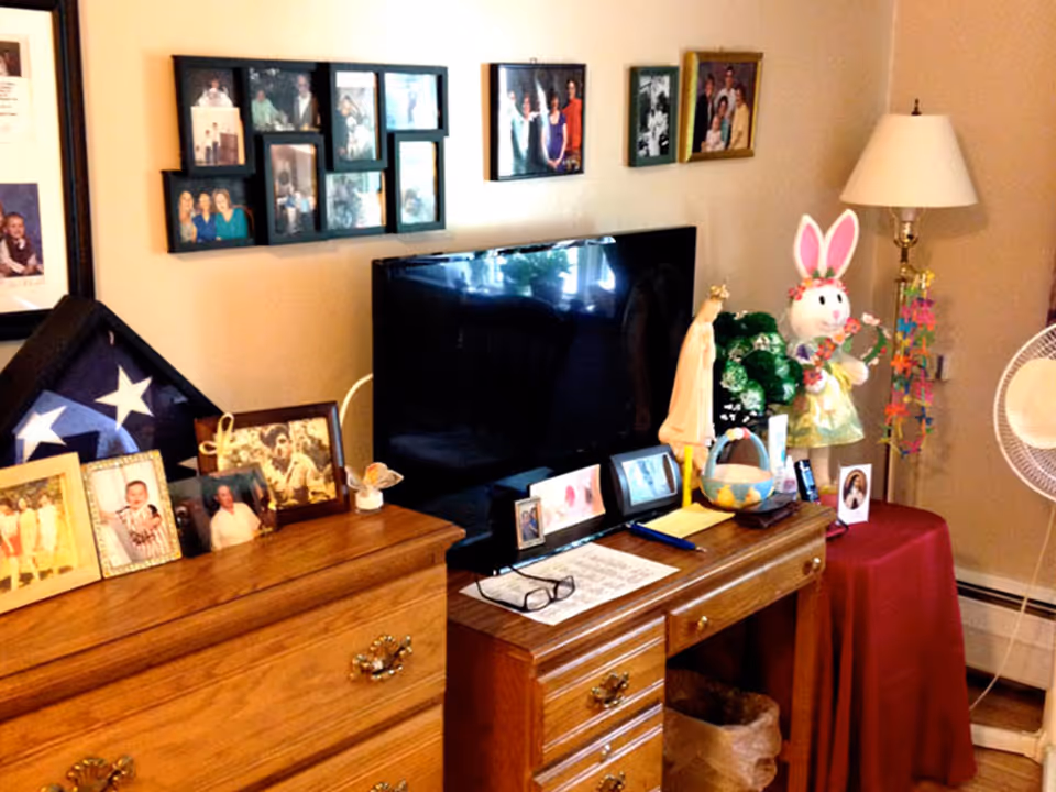 Wooden dresser and desk with a flat-screen TV, framed family photos, a lamp and decorative items including an Easter bunny in a cozy room.