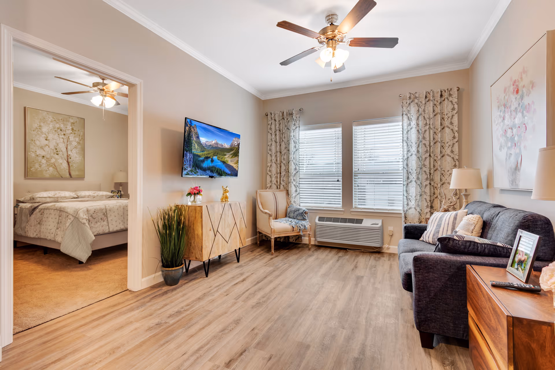 A bright and cozy living room with wood flooring, a ceiling fan, a wall-mounted TV, a decorative cabinet, an armchair with a throw blanket, and a dark gray sofa with pillows. The room has two large windows with patterned curtains and a wall-mounted air conditioning unit below them. To the left, an open doorway reveals a bedroom with a bed, nightstand, lamp, and a large floral painting on the wall.
