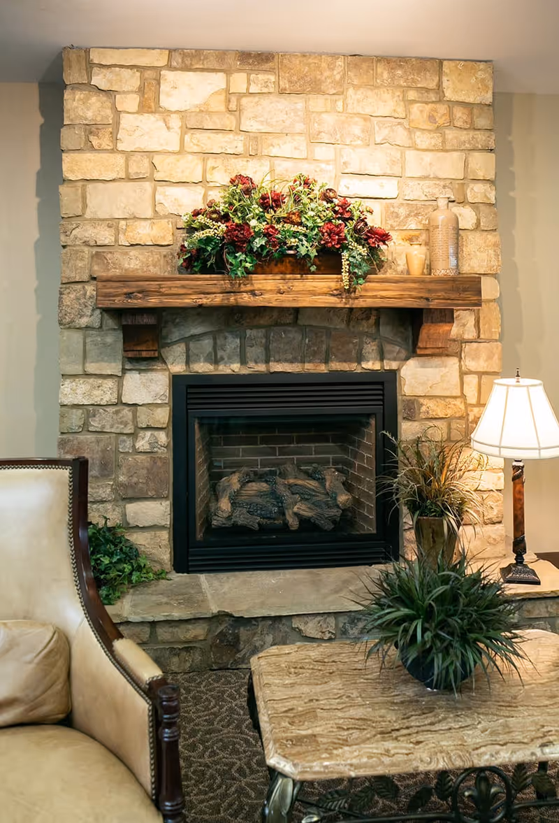 Stone fireplace with a wooden mantel decorated with a floral arrangement. In front of the fireplace is a beige armchair and a stone coffee table with a green potted plant. A table lamp with a white shade is on the right side on a small table.