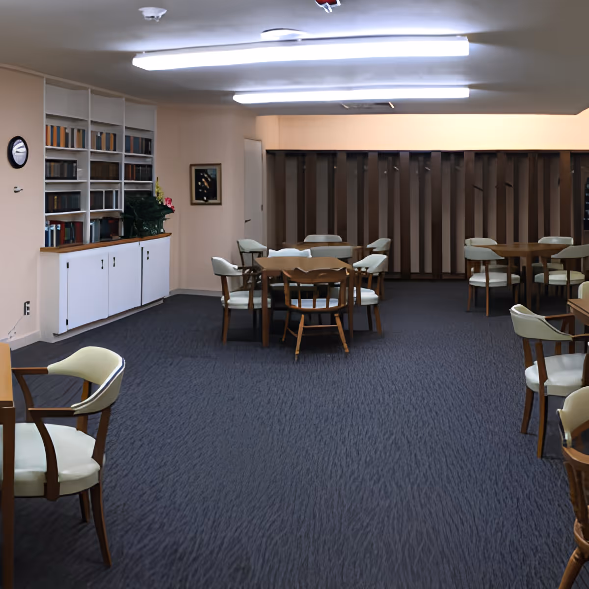 A spacious room with multiple wooden tables and cushioned chairs arranged on a dark carpeted floor. The walls are light-colored with built-in shelves filled with books and decorative plants. Fluorescent lights are mounted on the ceiling, illuminating the room evenly.