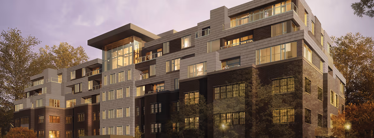 Exterior view of a modern multi-story senior living facility building at dusk with large windows and balconies, surrounded by trees with autumn foliage.