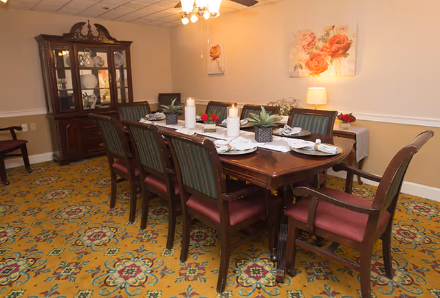 A formal dining room with a long wooden table set for eight people. The table is decorated with white candles, small potted plants, and neatly arranged plates with napkins. The room has a patterned yellow carpet, a wooden china cabinet with dishes, floral paintings on the wall, and a ceiling fan with lights.