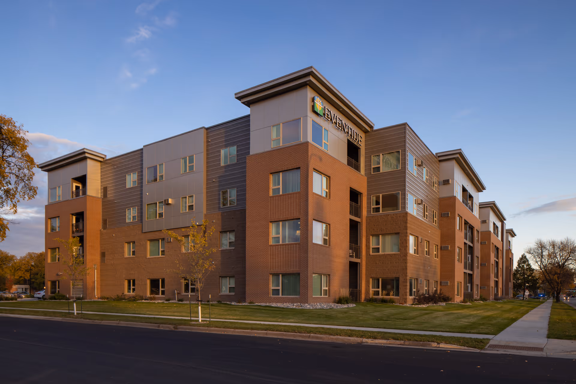 Four-story brick-and metal apartment building photographed from the street at dusk with a lawn and sidewalk in front.