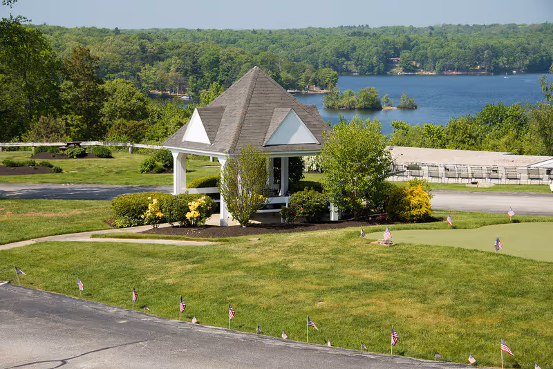 A small white gazebo with a gray roof surrounded by green bushes and trees, situated near a lake with a forested shoreline in the background. The foreground shows a well-maintained grassy area with small American flags placed along the edge of a paved surface.