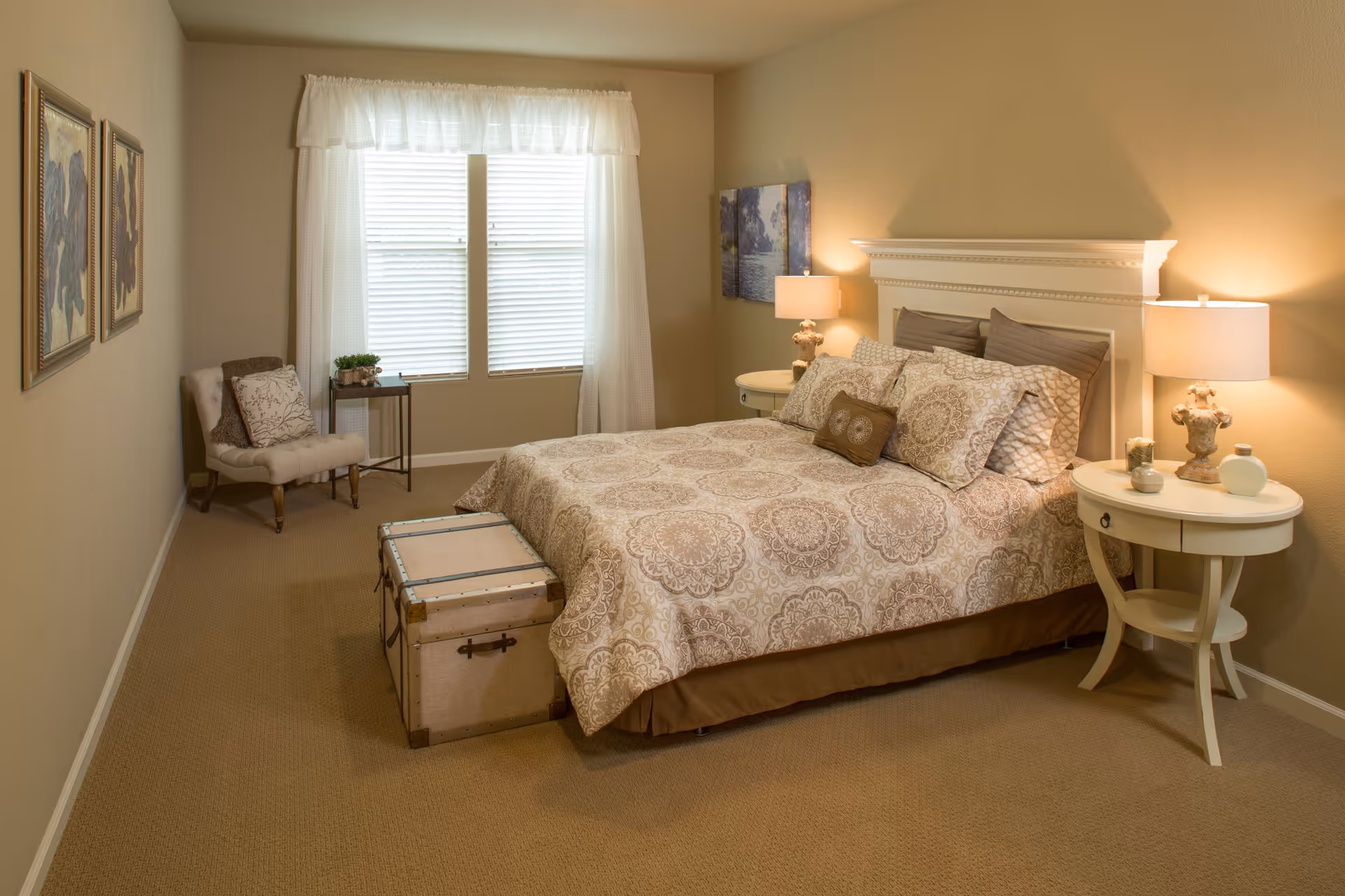 A cozy bedroom with a large bed covered in patterned bedding, two bedside tables with lamps, a cushioned chair with a pillow, two framed pictures on the wall, a window with white curtains, and a storage trunk at the foot of the bed.