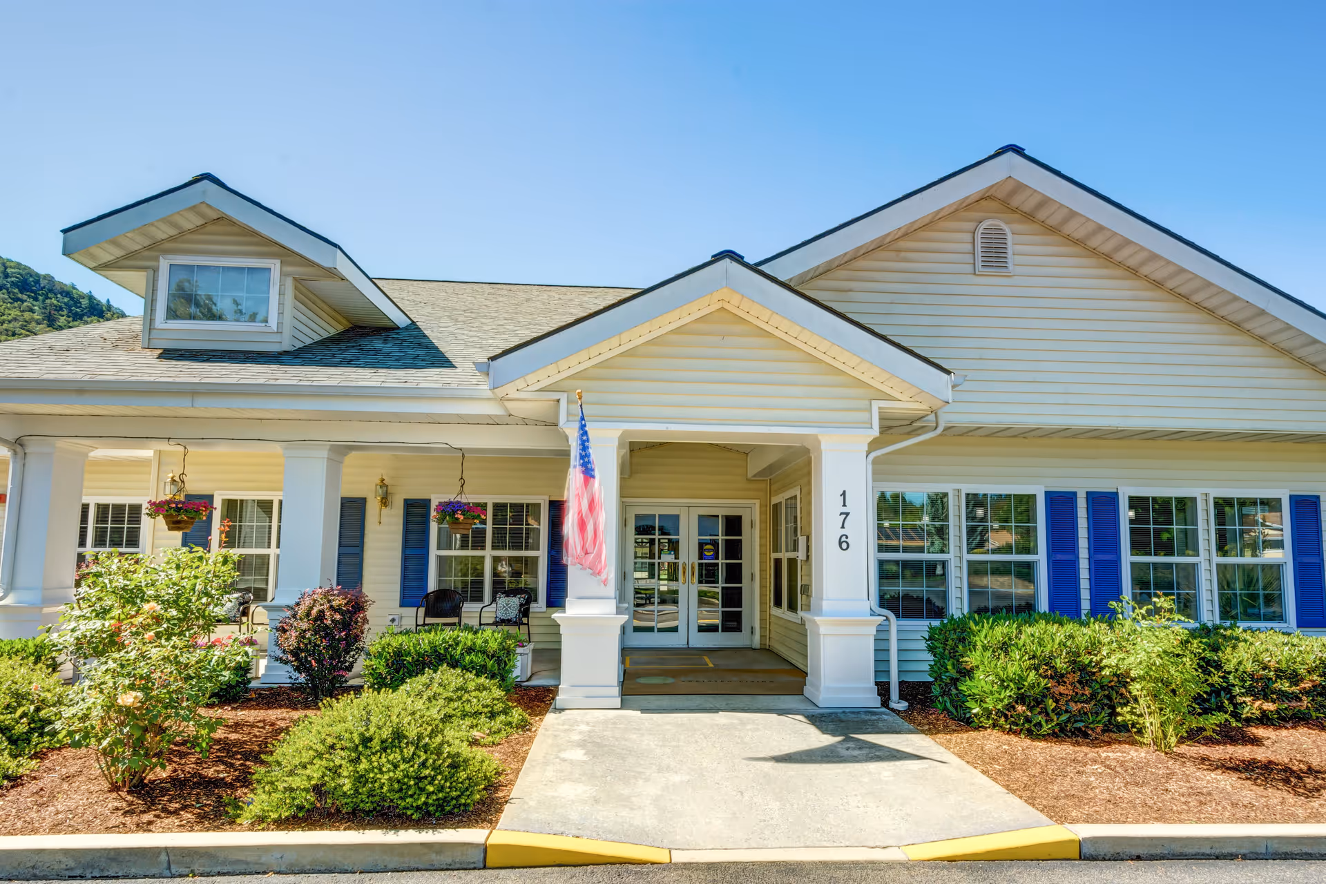 Front exterior view of Morrow Heights Assisted Living building with a covered entrance, white columns, blue shutters, hanging flower baskets, and an American flag. There are bushes and landscaped plants in front of the building under a clear blue sky.