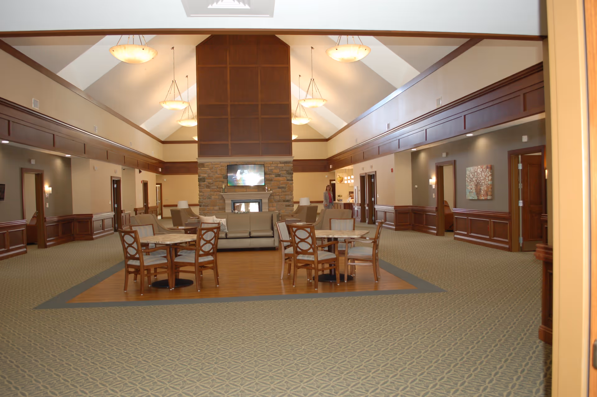 Spacious interior common area with high vaulted ceilings, multiple hanging light fixtures, and a central stone fireplace with a mounted TV above it. The room features several wooden tables with chairs and sofas arranged around the fireplace. The walls have wood paneling and neutral paint, with doorways leading to other rooms.