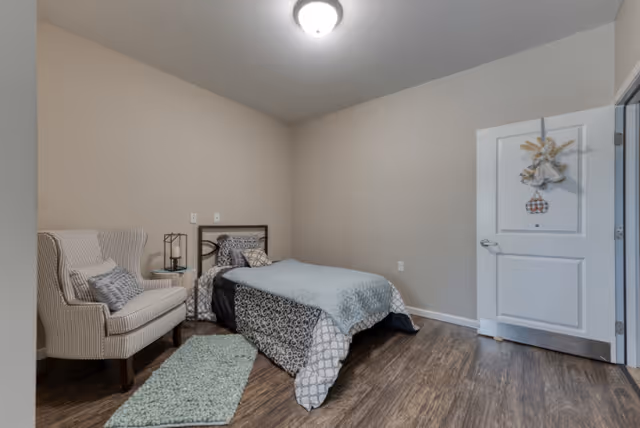 A simple bedroom with a single bed covered in patterned bedding and a light blue quilt. Next to the bed is a small round glass side table with a decorative lantern on it. A striped armchair with a cushion is placed near the bed. The room has wooden flooring, beige walls, a white door with a decorative hanging, and a ceiling light fixture.