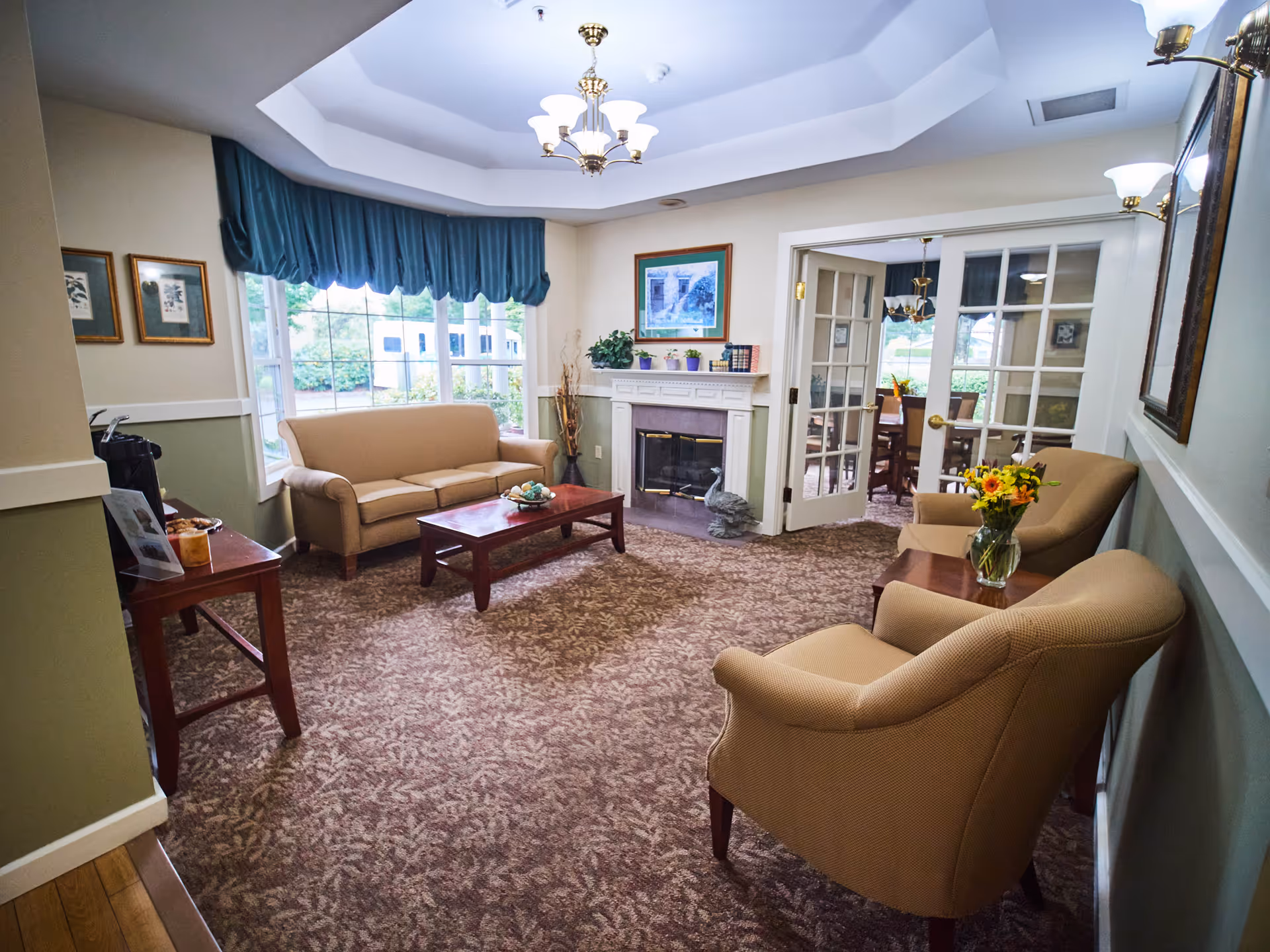 A cozy senior living facility common area with beige upholstered chairs and a sofa arranged around a wooden coffee table. The room features a fireplace with decorative items on the mantel, framed artwork on the walls, and large windows with blue valances letting in natural light. A glass door leads to a dining area with a table and chairs. The carpet has a floral pattern and the walls are painted in soft green and cream tones.