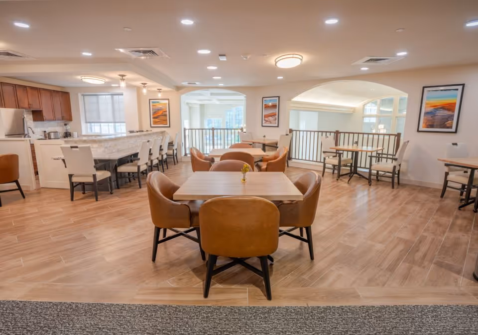 A spacious dining area in a senior living facility featuring several tables with brown leather chairs. The room has wood flooring, a kitchen area with a marble countertop and bar stools, and framed artwork on the walls. Large windows allow natural light to brighten the space.
