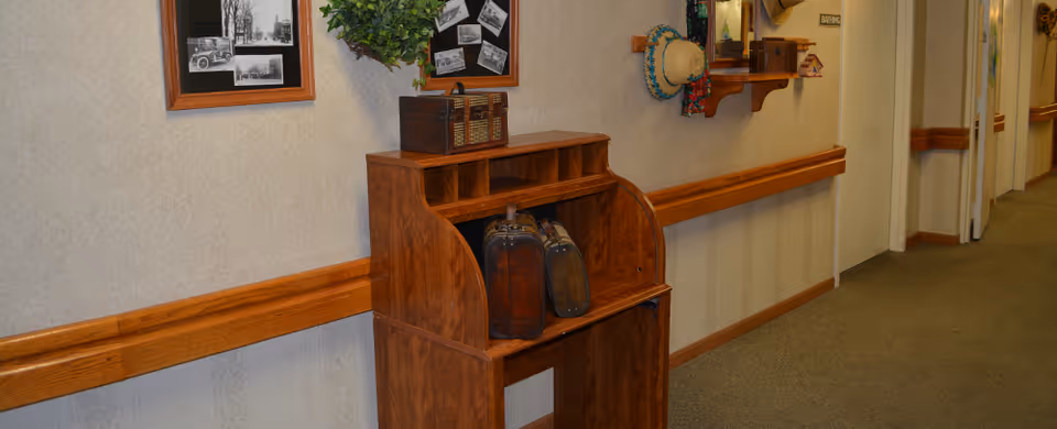 A hallway in a senior living facility with beige walls and carpeted floor. There is a wooden roll-top desk against the wall with vintage suitcases on it. Above the desk are framed black and white photos and a small green plant. Further down the hallway, there is a wooden shelf with a mirror and a hat hanging on the wall.