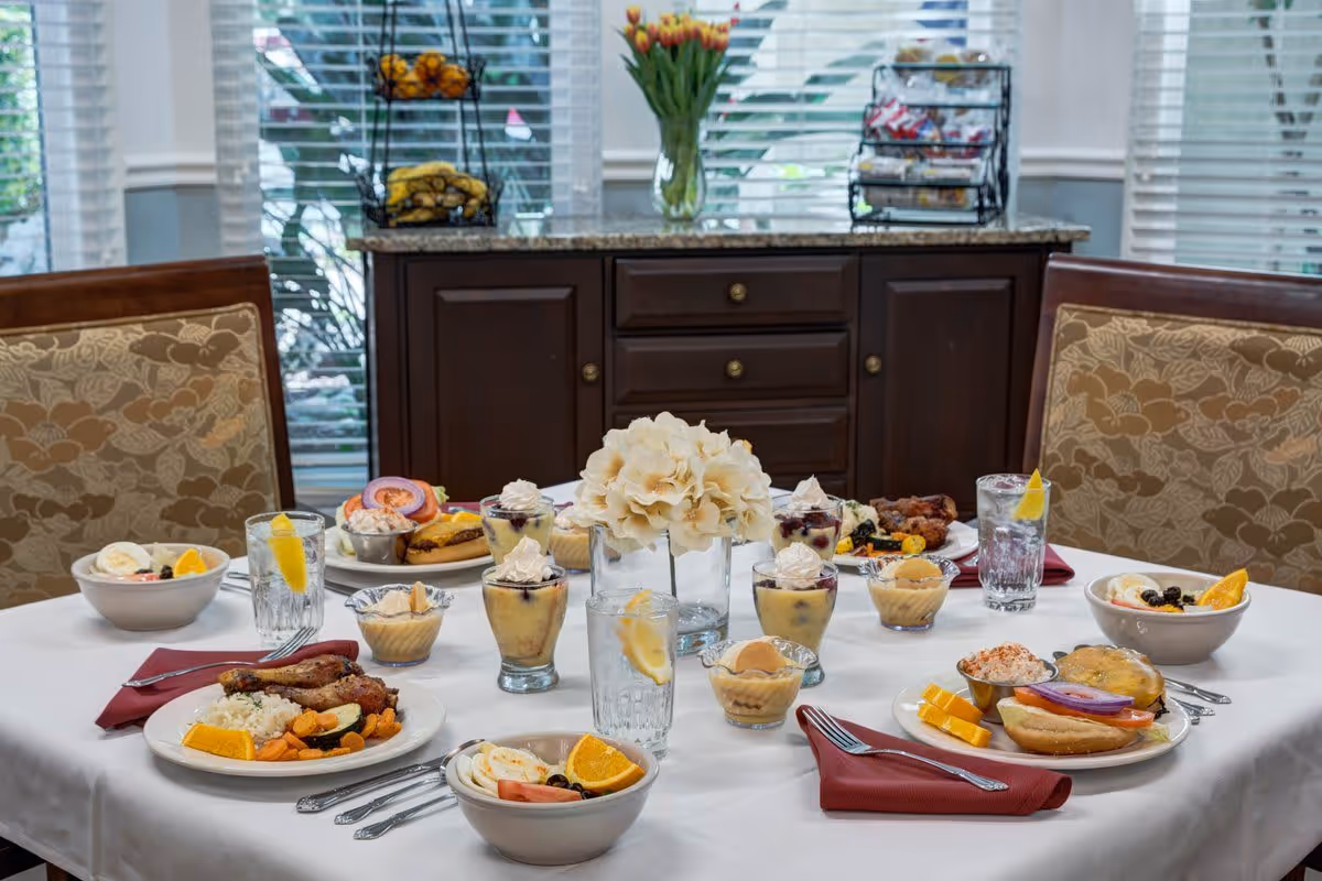 A dining table set with various plated meals including meat, vegetables, fruit, and desserts, along with glasses of water with lemon slices. The table is covered with a white tablecloth and has a centerpiece of white flowers. Behind the table is a wooden cabinet with snacks and a vase of tulips, and large windows with blinds letting in natural light.