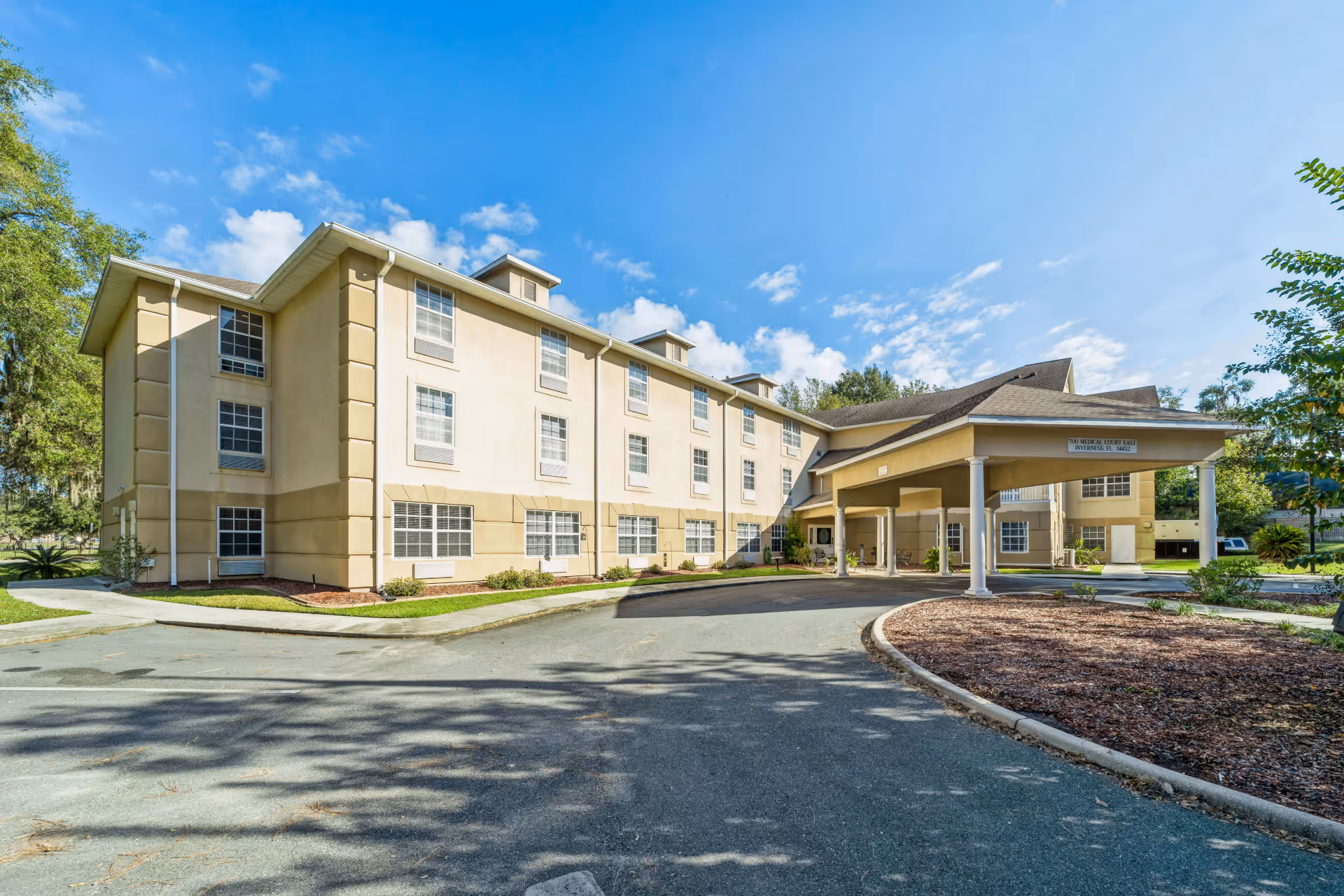 Exterior view of a three-story beige senior living facility building with multiple windows and a covered entrance driveway under a clear blue sky.