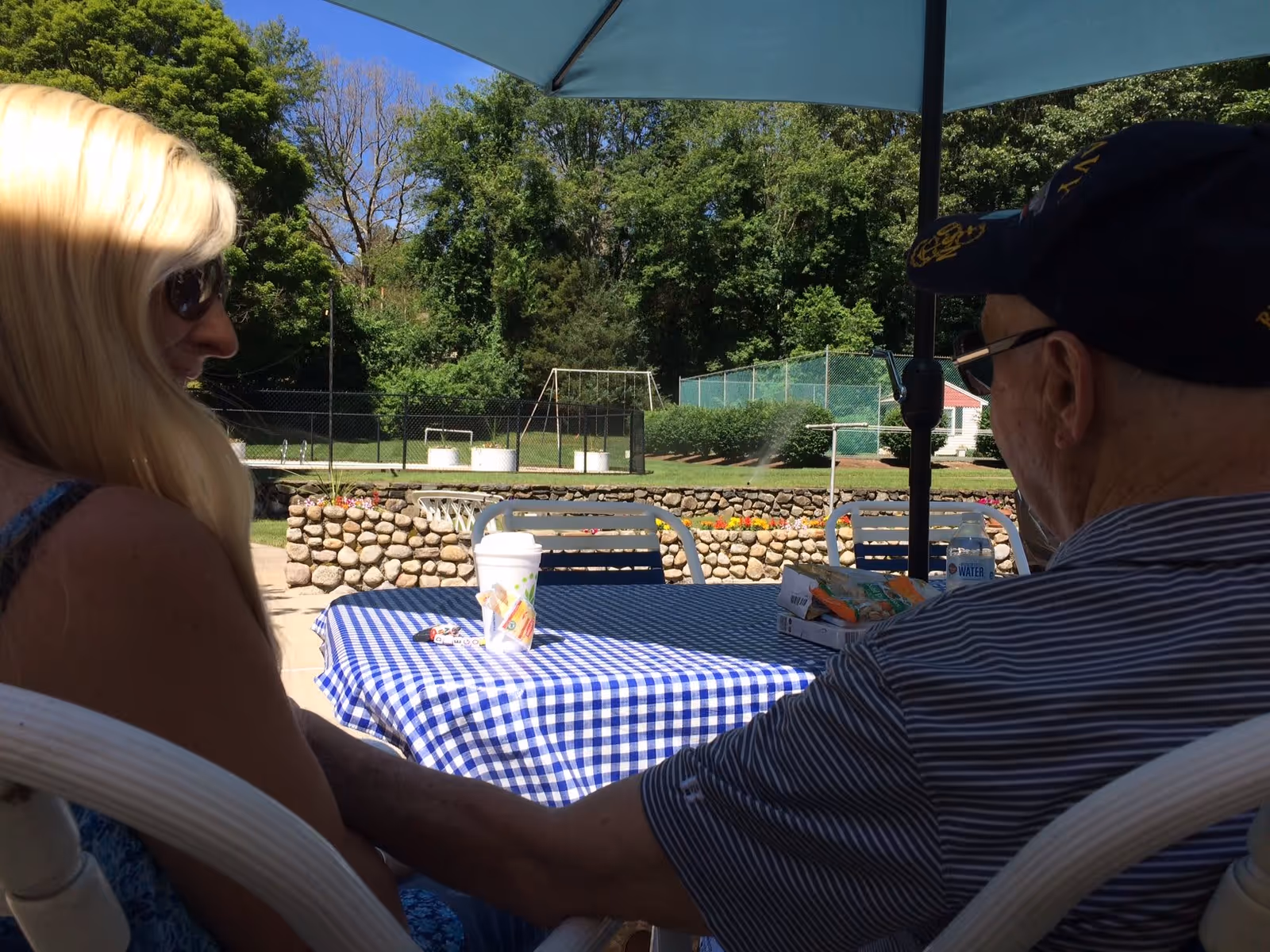 Two people sit at a blue-checked outdoor table under an umbrella with a stone wall and tennis courts in the background.