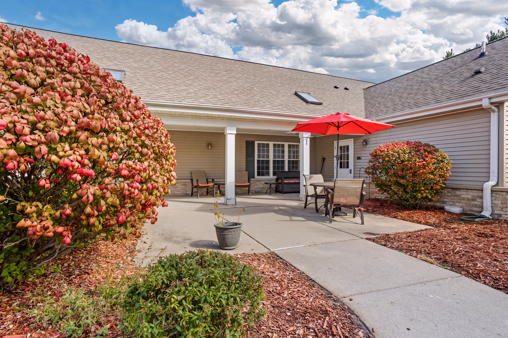 Outdoor patio area at Brookdale Delta with a concrete walkway, red and green bushes, a table with four chairs, and a red umbrella. The building has beige siding and a sloped roof under a partly cloudy sky.