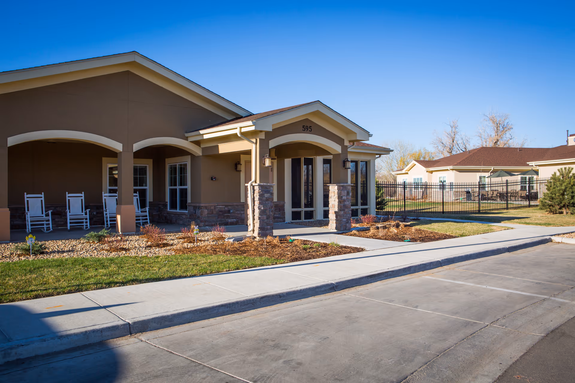 Front entrance of a senior living building with a covered porch, rocking chairs, and landscaped walkway.