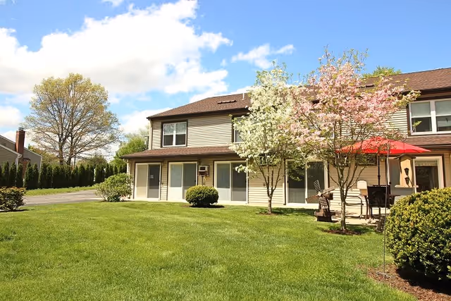 Exterior view of a two-story residential building with beige siding and brown roof. The foreground features a well-maintained green lawn, flowering trees with white and pink blossoms, and a patio area with outdoor furniture and a red umbrella. The sky is partly cloudy with blue patches.