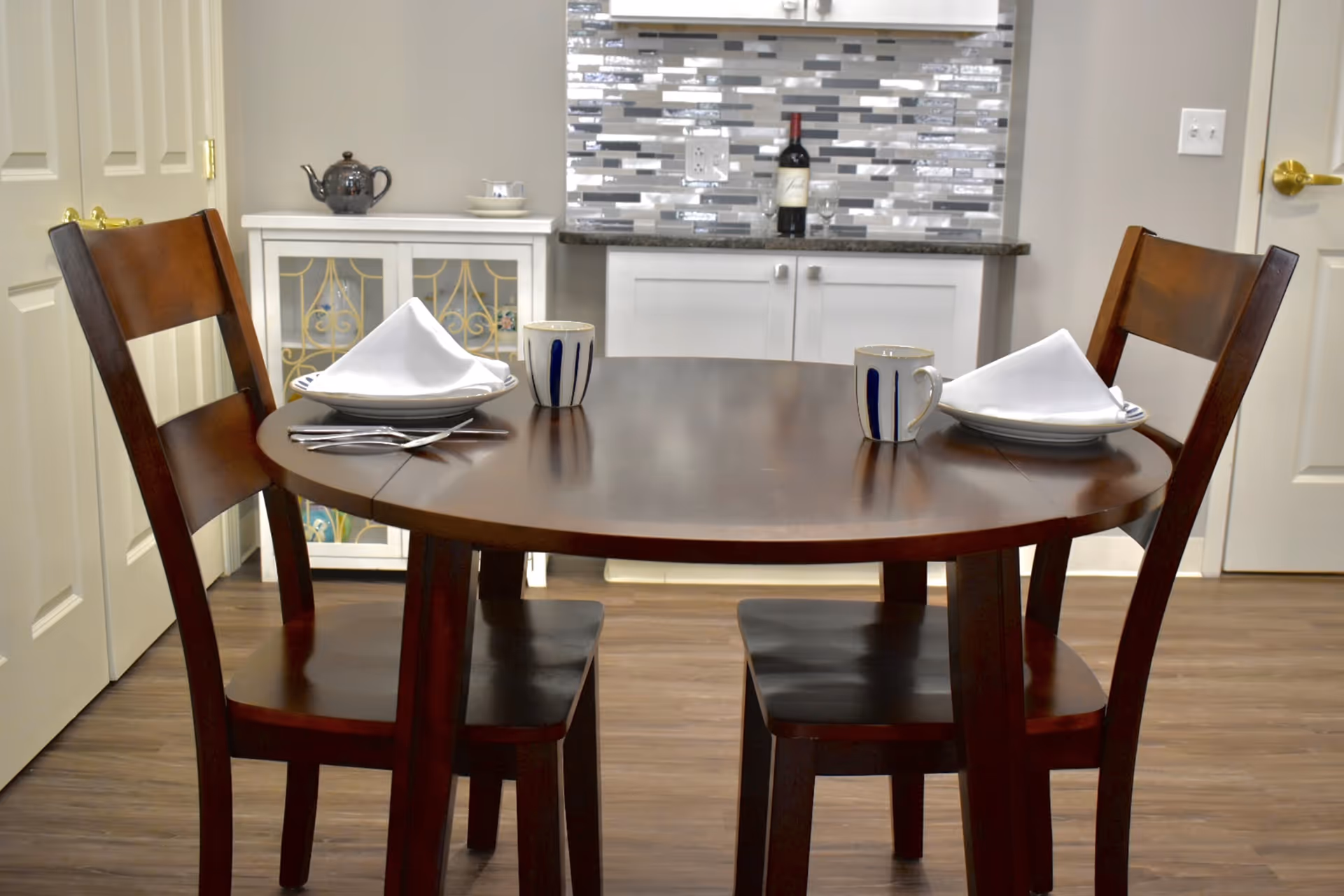 A small dining area with a round wooden table set for two, featuring white plates with folded white napkins, striped mugs, and silverware. Behind the table is a cabinet with decorative glass doors and a countertop with a bottle of wine and two wine glasses. The backsplash is made of rectangular tiles in shades of gray and white.