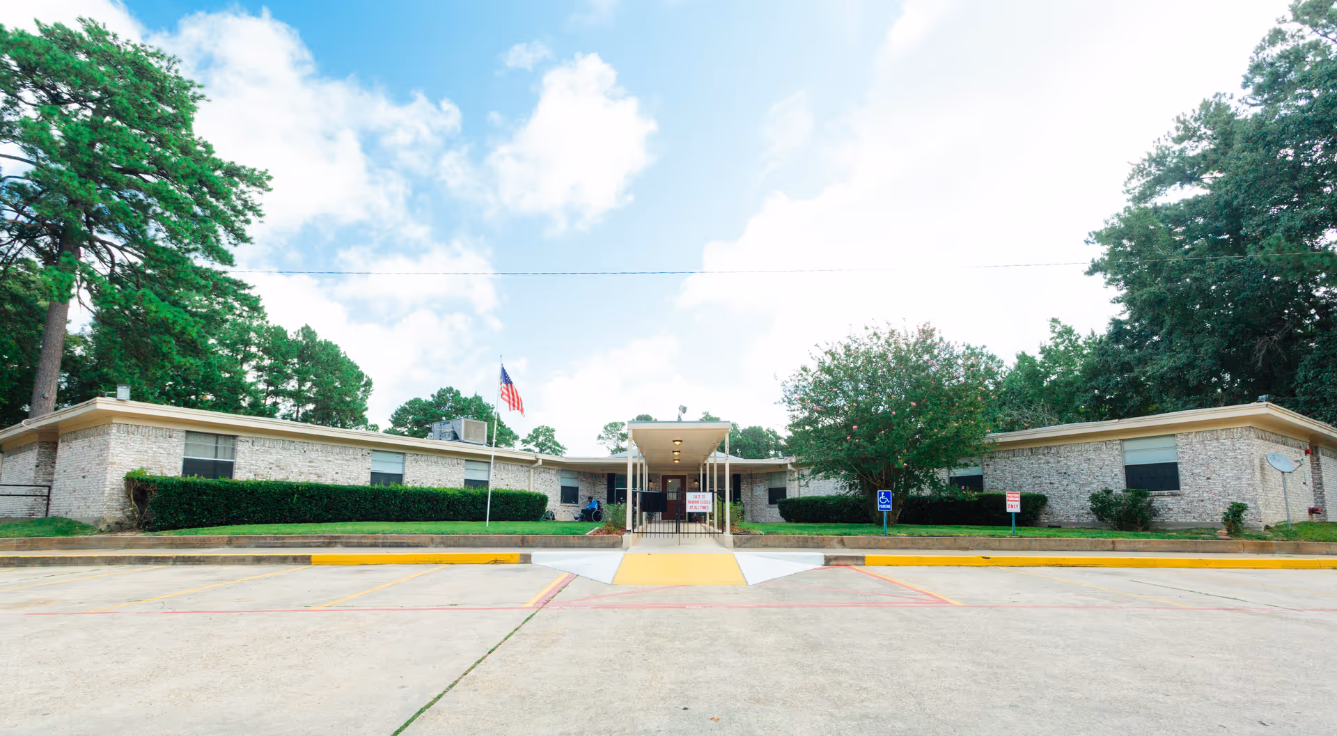 Front exterior view of a single-story brick building with a covered entrance, an American flag on a flagpole, and surrounding greenery including trees and bushes under a partly cloudy sky.