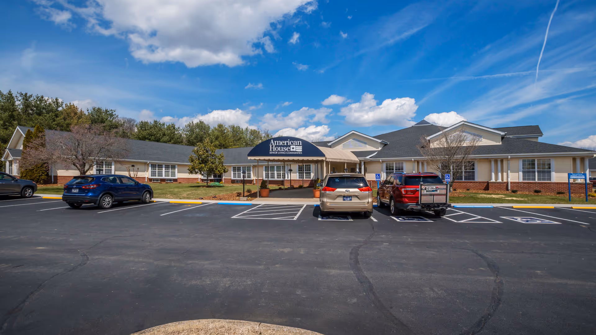 Front exterior view of American House Johnson City senior living community building with a covered entrance, several parked cars, and a clear blue sky with some clouds.