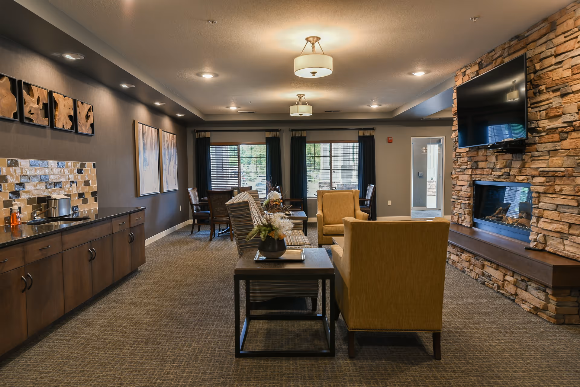 A cozy living room area in a senior living facility with comfortable armchairs and a sofa arranged around a coffee table. The room features a stone fireplace with a mounted flat-screen TV above it. There are large windows with dark curtains letting in natural light, and a sideboard with a sink and decorative backsplash along one wall. The ceiling has recessed lighting and two hanging light fixtures.