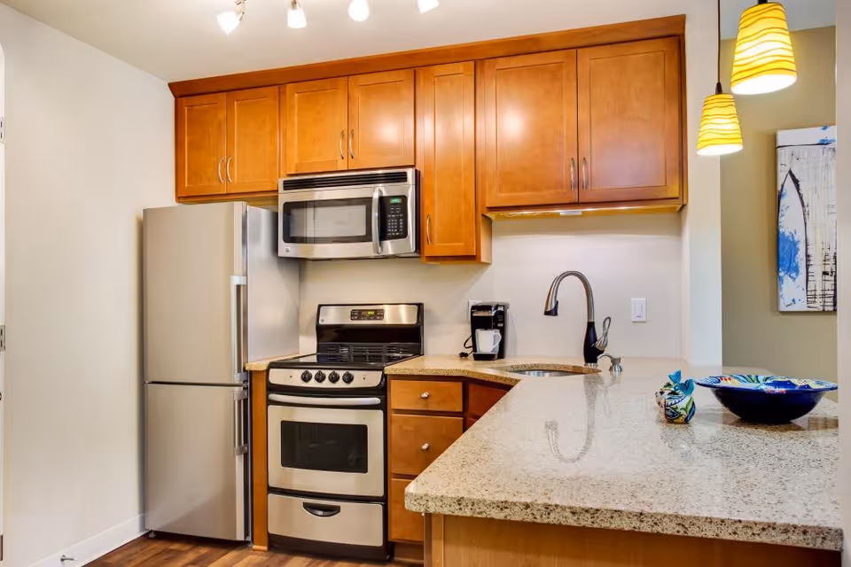 A modern kitchen with wooden cabinets, a stainless steel refrigerator, a microwave mounted above a stove, a coffee maker on the counter, and a granite countertop with a sink and two hanging pendant lights above it. There is also a decorative bowl and a small colorful item on the countertop.