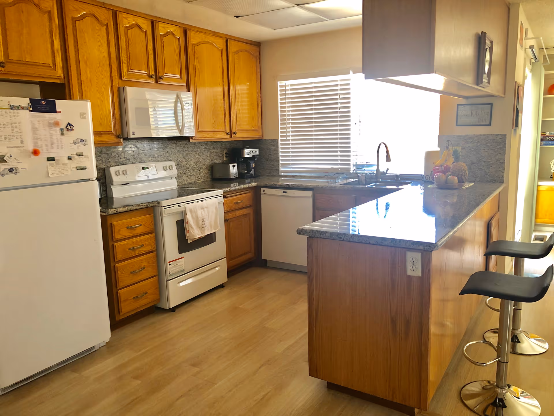 A bright kitchen with wooden cabinets, a white refrigerator covered with papers and magnets, a white stove with a towel hanging on the handle, a microwave above the stove, a dishwasher, and a granite countertop island with two black bar stools. A window with blinds is above the sink, and there is a bowl of fruit on the counter.