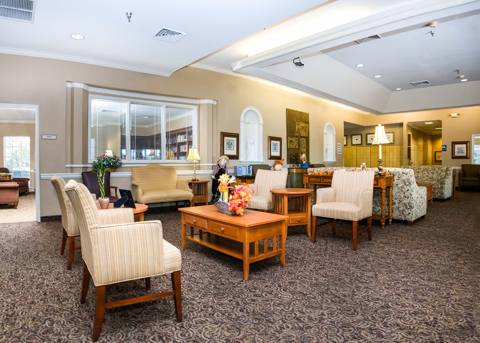 A spacious and well-lit senior living facility common area with multiple seating arrangements including striped armchairs, a beige loveseat, and floral-patterned sofas. Wooden coffee tables with decorative flower arrangements are placed in the center. The walls are adorned with framed artwork and niches containing statues. The carpet has a patterned design, and there are several table lamps providing warm lighting. A hallway and additional seating areas are visible in the background.