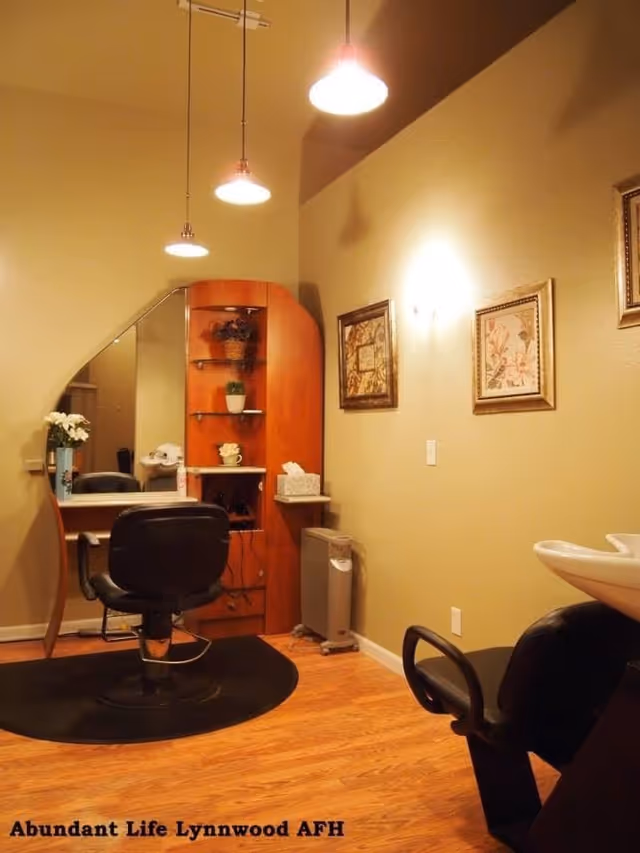 A warm-lit interior salon area with a styling chair in front of a mirror and wooden shelving, a wash basin to the right, and framed artwork on the wall.