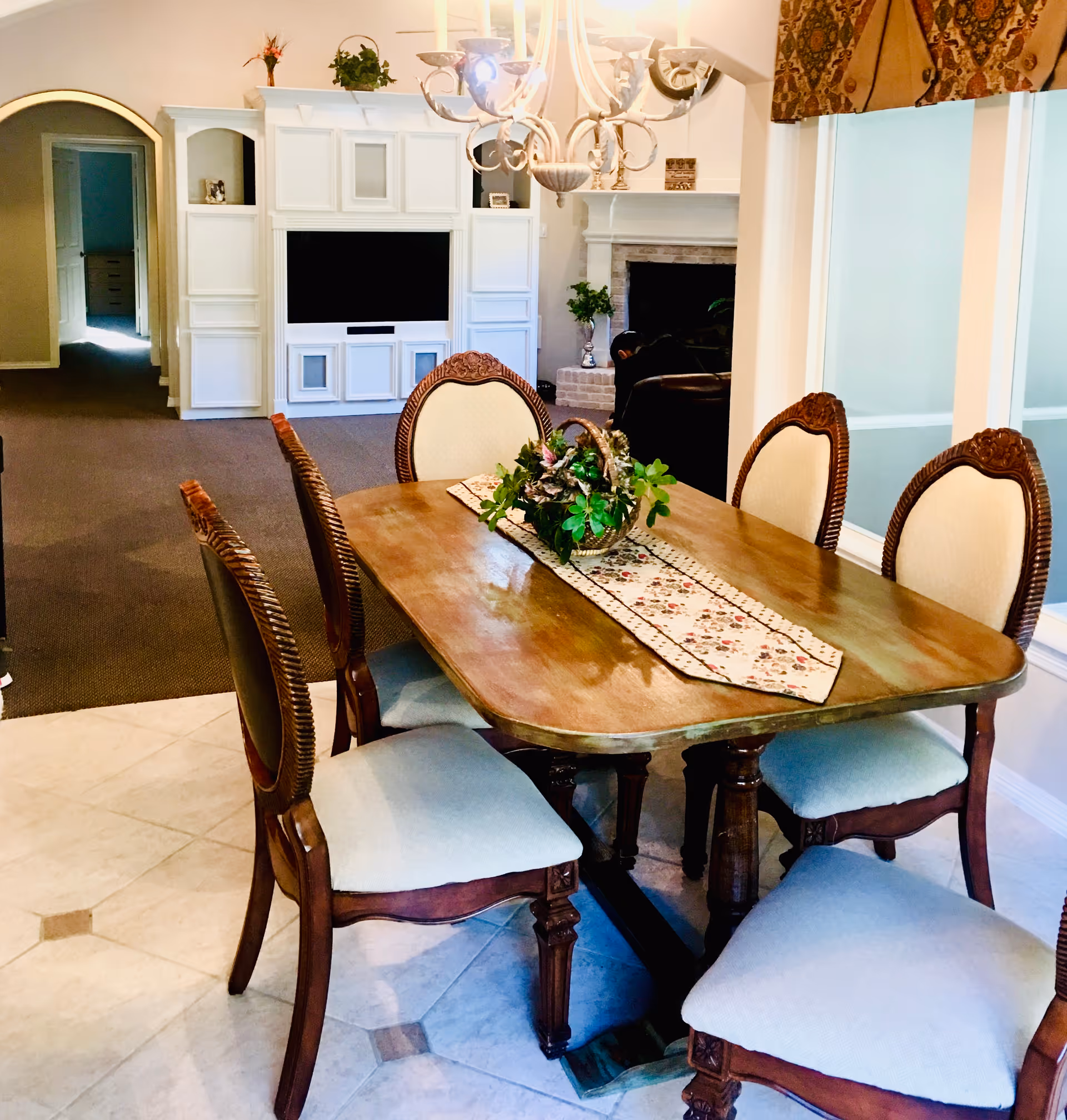 A dining area with a wooden table and six upholstered chairs. The table has a floral table runner and a decorative basket with greenery. In the background, there is a white entertainment center with a TV, a fireplace, and an arched doorway leading to another room.