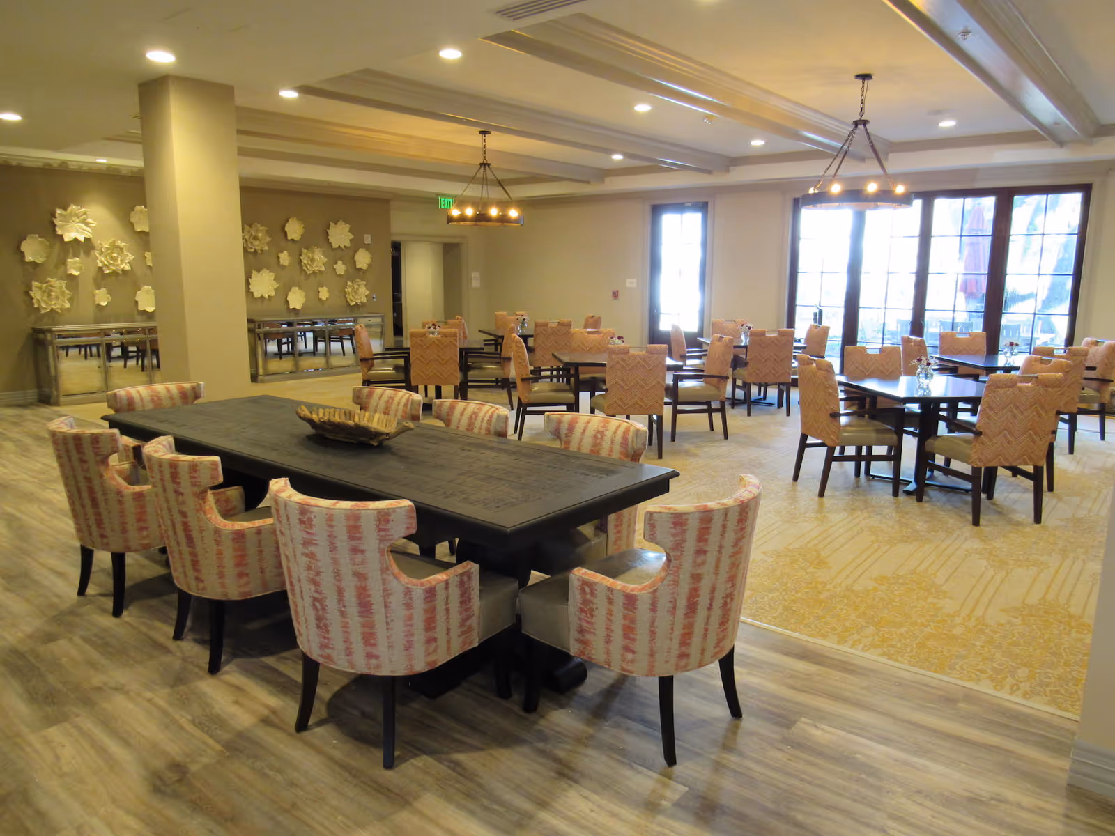 A spacious dining room with multiple tables and cushioned chairs arranged neatly. The room features large windows allowing natural light to enter, decorative wall art with floral designs, and warm lighting fixtures hanging from the ceiling. The floor has a combination of wood and carpeted areas.