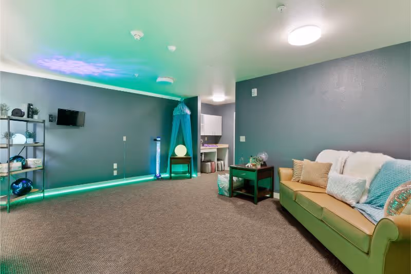 A cozy room with gray walls and carpeted floor featuring a green sofa with multiple pillows and a throw blanket. There is a small dark wooden side table next to the sofa with decorative items on it. On the left side of the room, there is a black metal shelving unit with various decorative objects. The room has ambient green and purple lighting along the ceiling and floor edges, creating a calming atmosphere. In the background, there is a small alcove with white cabinets and storage baskets.