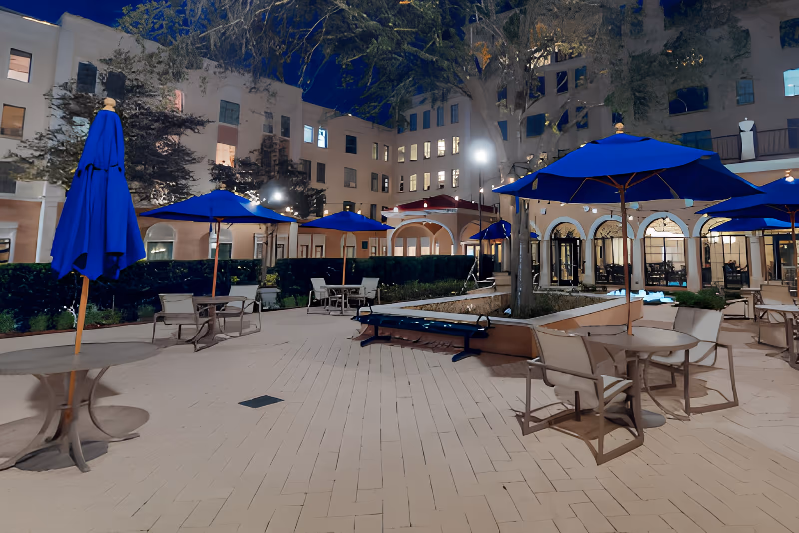 Outdoor courtyard area at night with multiple tables and chairs under blue umbrellas. The courtyard is surrounded by a multi-story building with lit windows and some trees. There are benches and planters integrated into the paved area.
