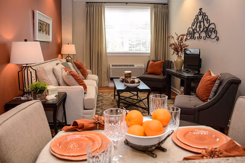 A cozy living room and dining area with a beige sofa adorned with orange and beige pillows, two gray armchairs with orange pillows, a black coffee table with decorative items, a side table with a lamp and plant, and a dining table set with orange plates, crystal glasses, and a bowl of oranges. The room has a window with beige curtains and a decorative metal wall hanging above a small TV on a dark wooden table.