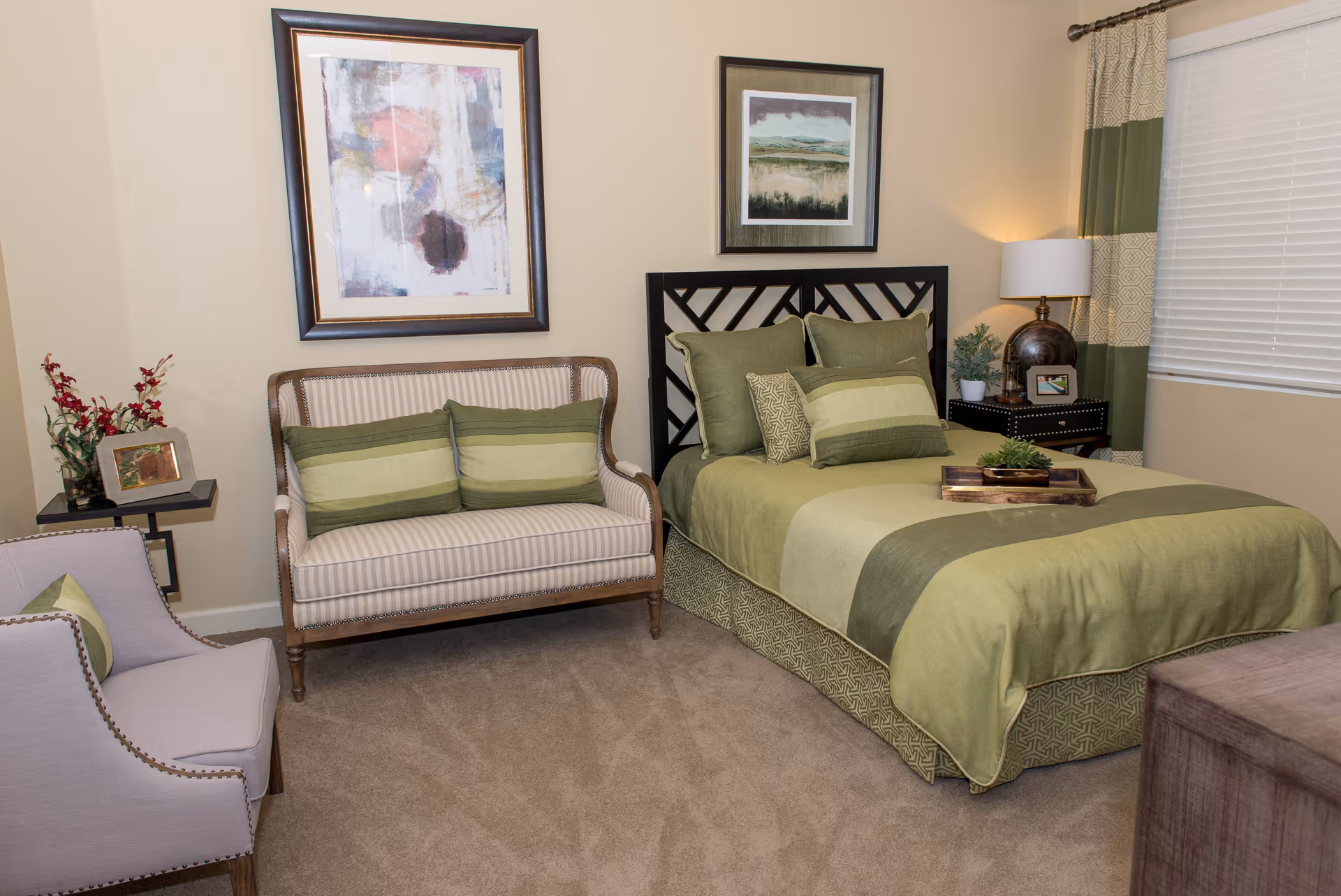 A cozy bedroom featuring a bed with green and beige bedding, a wooden headboard, and multiple pillows. Next to the bed is a nightstand with a lamp, a small plant, and a framed photo. There is a striped loveseat with green pillows and a light purple armchair with a green pillow. Two framed artworks hang on the beige walls, and a window with green and beige curtains is visible.