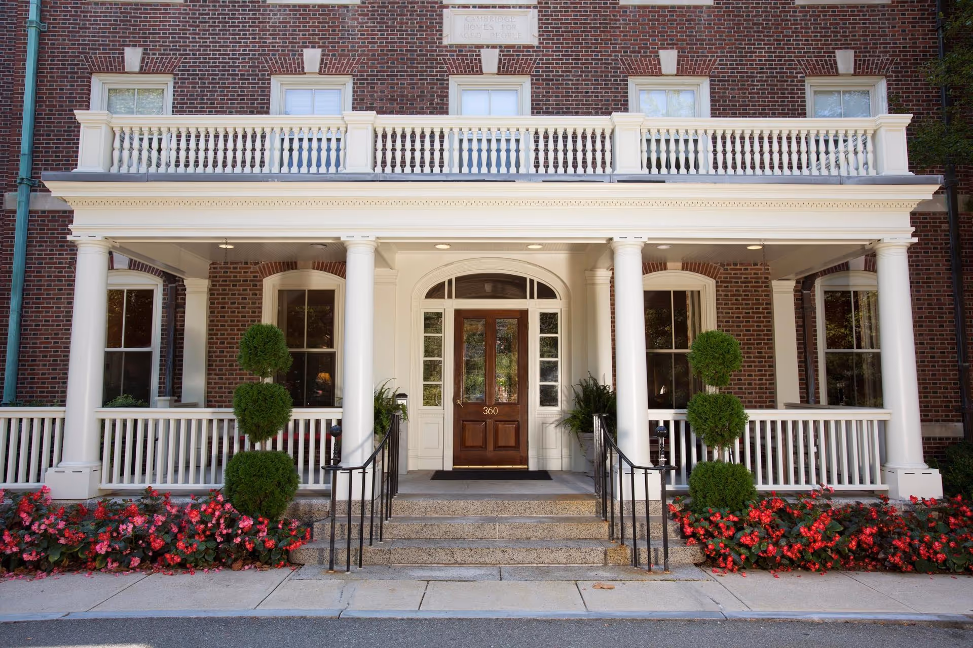 Front entrance of The Cambridge Homes building featuring a brick facade, white columns, a wooden door with the number 360, decorative topiary plants, and flower beds with red flowers along the walkway.