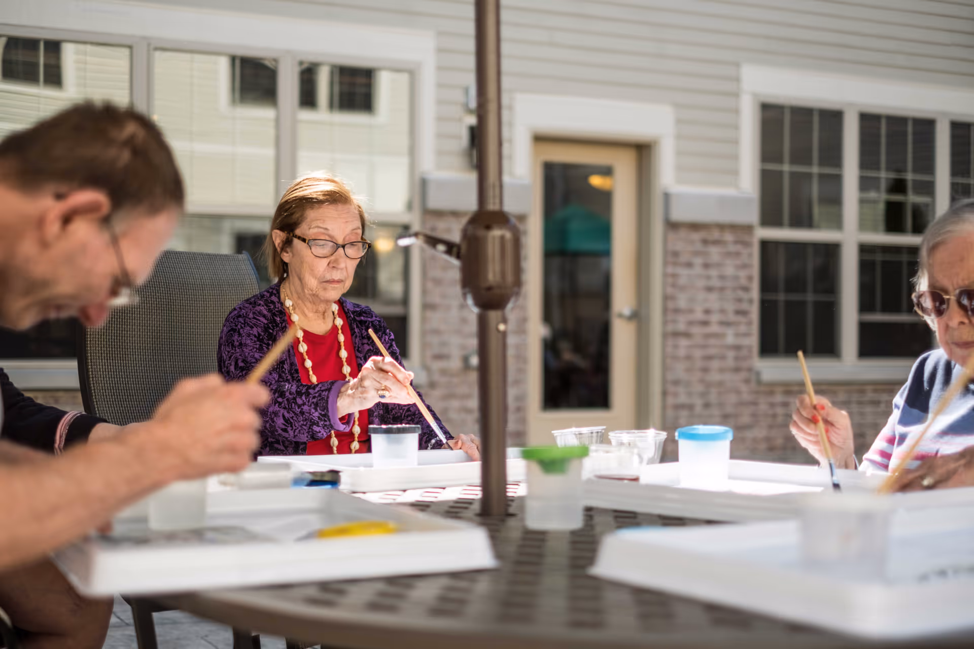 Three elderly individuals sitting around a round outdoor table engaged in painting activities with brushes and paint containers, with a building featuring windows and a door in the background.