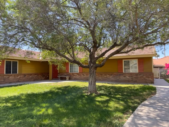 Single-story building with a yellow and brick exterior, red shutters, and a red door, surrounded by a green lawn and a large tree in the foreground under a clear blue sky.
