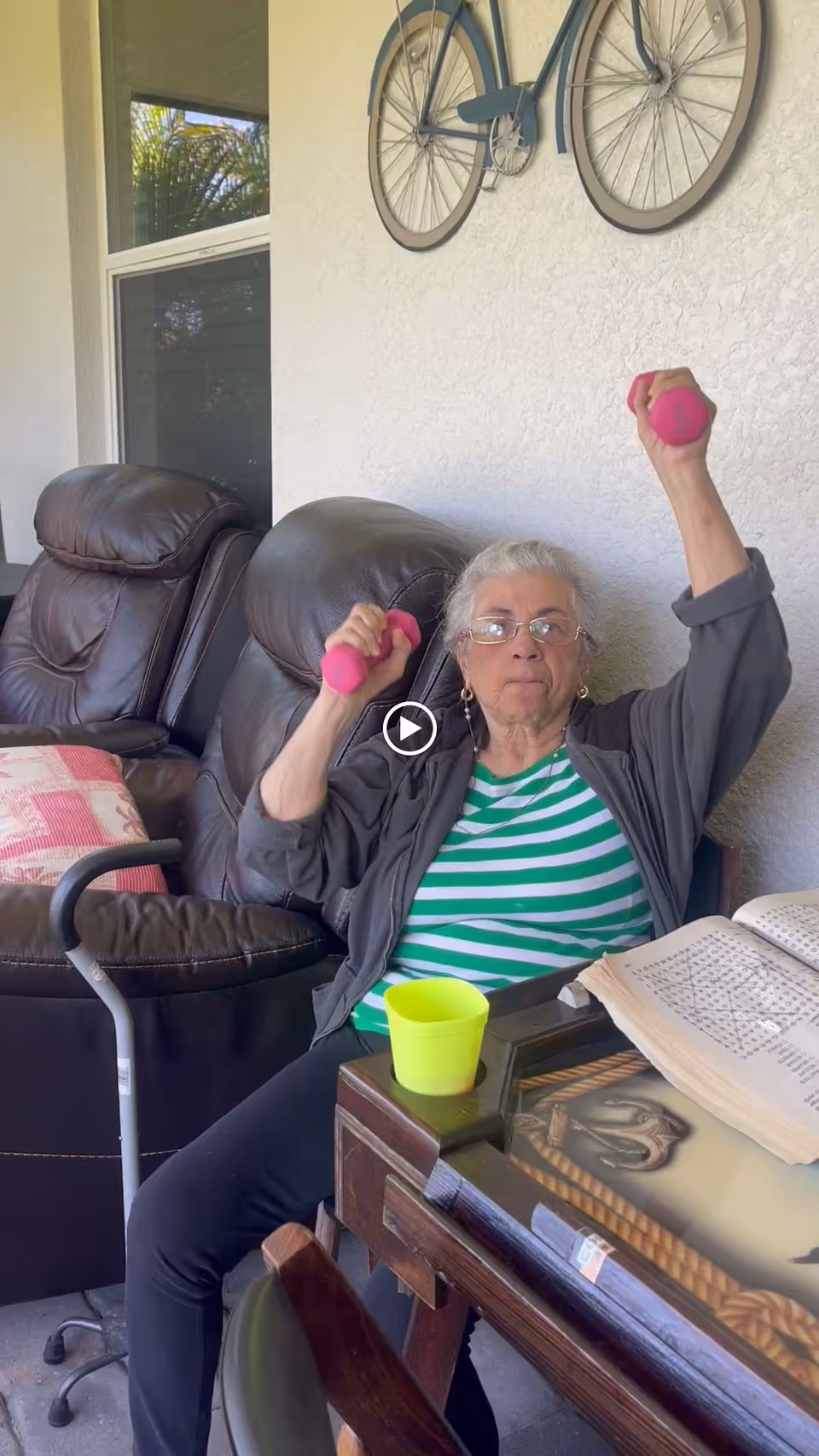 An elderly woman wearing glasses and a green and white striped shirt is sitting in a chair on a covered patio, lifting pink hand weights. Behind her are two brown leather recliners and a wall with a decorative bicycle mounted on it. A walking cane rests against one recliner, and a table with a yellow cup and an open book is in front of her.
