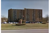 A multi-story brown residential building with balconies on each floor, surrounded by a parking lot with several cars and a grassy area with a small hedge and flagpole in front.