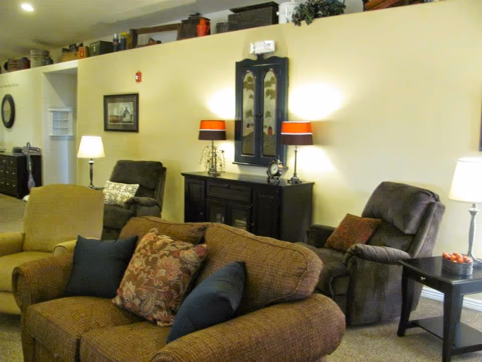 A cozy living room area with a brown sofa adorned with patterned and solid pillows, a beige armchair, and two dark recliners. The walls are light yellow with framed artwork and a decorative cabinet with two lamps on top. There are side tables with lamps and decorative items, and the room has a warm, inviting atmosphere.