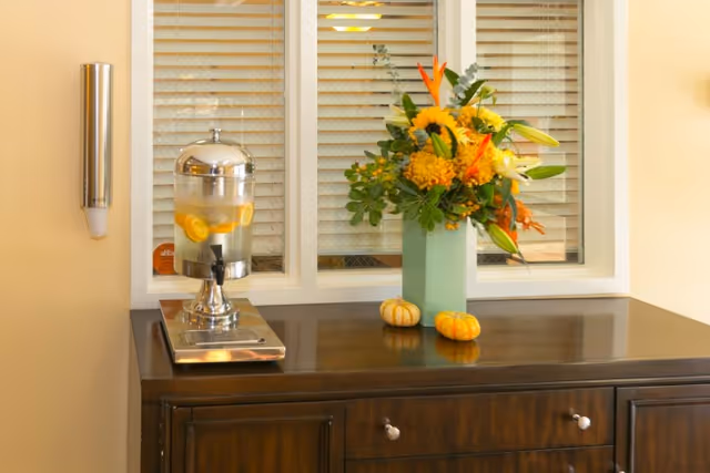 A dark wooden cabinet with a glass dispenser filled with lemon water on the left side and a green vase with a colorful flower arrangement on the right side. Two small decorative pumpkins are placed in front of the vase. Behind the cabinet is a window with closed blinds.