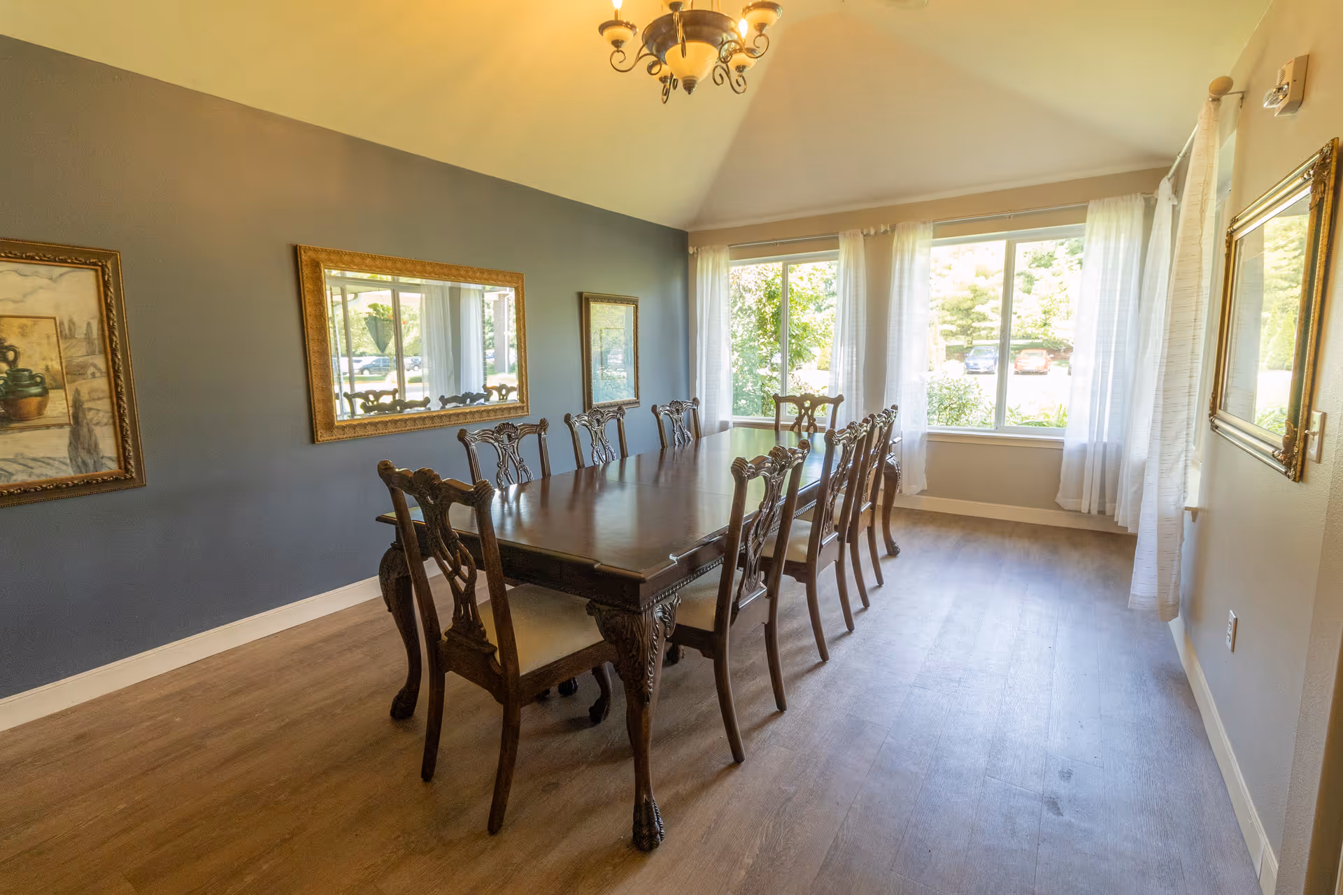 Bright dining room with a long wooden table surrounded by ornate chairs, large windows with sheer curtains, and framed mirrors and artwork on the walls.